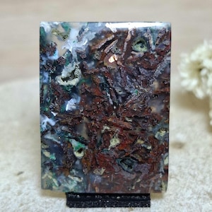 May include: A rectangular gemstone with a complex pattern of brown, green, and white inclusions. The stone is mounted on a black stand, showcasing its natural textures and colors. The overall effect is a unique, earthy aesthetic, perfect for jewelry or display.