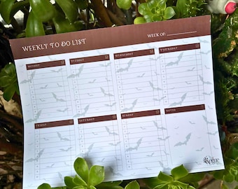 May include: A weekly to-do list notepad with a brown header and bat illustrations. The notepad is designed with sections for each day of the week, with space for notes. The notepad is surrounded by green plants.