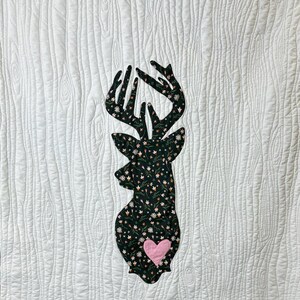 May include: White quilted wall hanging with a deer silhouette. The deer is filled with a floral pattern and a pink heart. The edges are trimmed with a floral pattern. The wall hanging is hung on a metal rod with wooden clothespins.