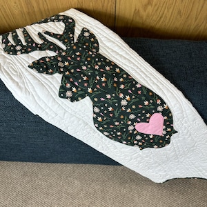 May include: White quilted throw blanket featuring a deer head silhouette. The deer head is made of dark green fabric with a floral pattern and a pink heart. The blanket is on a blue sofa.