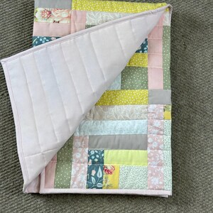 May include: A patchwork quilt with a light pink backing and a variety of patterned squares in pink, green, yellow, and gray. The quilt is folded over, revealing the quilted backing.