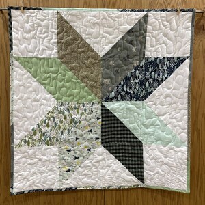 May include: A quilted star wall hanging with a variety of fabrics in shades of green, gray, and white. The star is centered on a white background with quilted texture. The quilt is hung on a wooden dowel with clothespins.