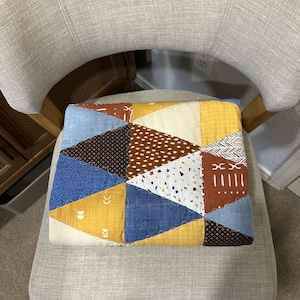 Puede incluir: Un coj&iacute;n de patchwork con un dise&ntilde;o geom&eacute;trico de tri&aacute;ngulos en varios colores, incluyendo azul, amarillo, marr&oacute;n y blanco. El coj&iacute;n est&aacute; colocado sobre una silla gris tapizada.