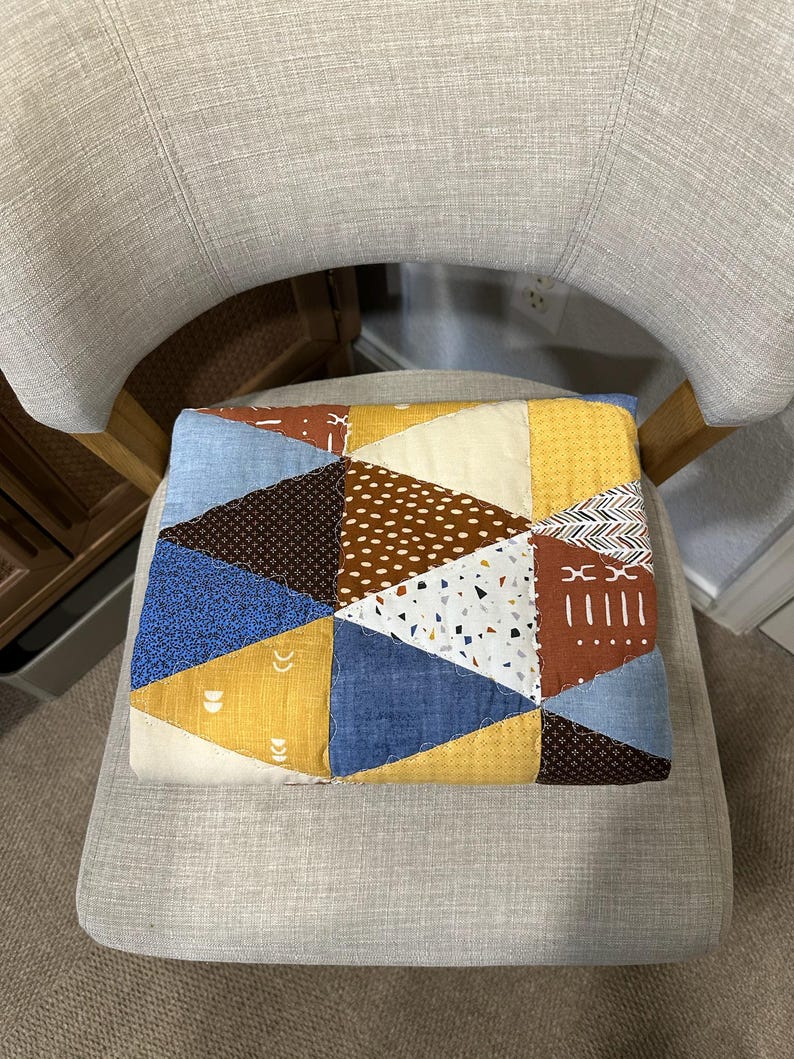 Puede incluir: Un coj&iacute;n de patchwork con un dise&ntilde;o geom&eacute;trico de tri&aacute;ngulos en varios colores, incluyendo azul, amarillo, marr&oacute;n y blanco. El coj&iacute;n est&aacute; colocado sobre una silla gris tapizada.