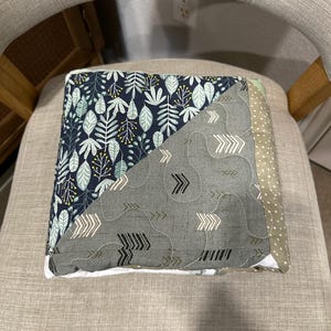 May include: A folded patchwork quilt with a botanical and arrow pattern. The quilt features a navy blue fabric with white and green leaves, a gray fabric with white and black arrows, and a green polka dot fabric. The quilt is on a gray chair.