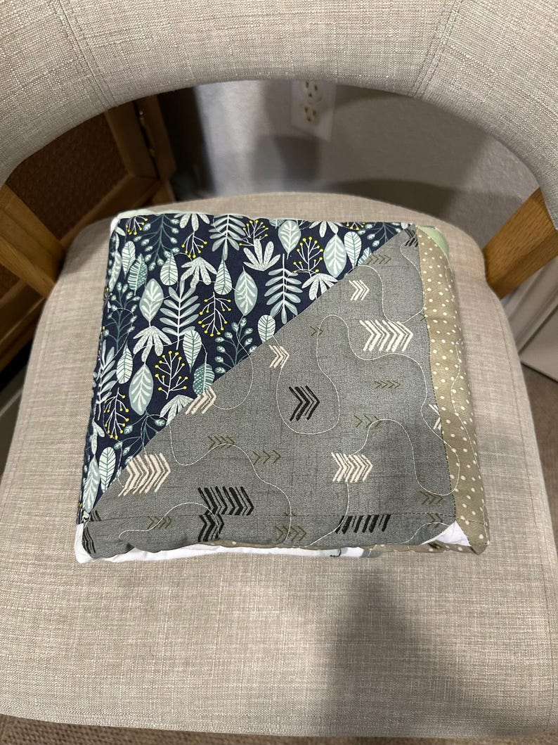May include: A folded patchwork quilt with a botanical and arrow pattern. The quilt features a navy blue fabric with white and green leaves, a gray fabric with white and black arrows, and a green polka dot fabric. The quilt is on a gray chair.