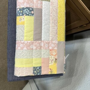 May include: A patchwork quilt with a variety of floral and geometric patterns in pastel colors, including pink, yellow, and blue. The quilt is made of squares and rectangles of fabric, and is placed on a blue upholstered bench.