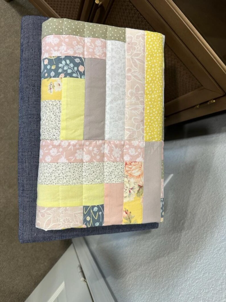 May include: A patchwork quilt with a variety of floral and geometric patterns in pastel colors, including pink, yellow, and blue. The quilt is made of squares and rectangles of fabric, and is placed on a blue upholstered bench.