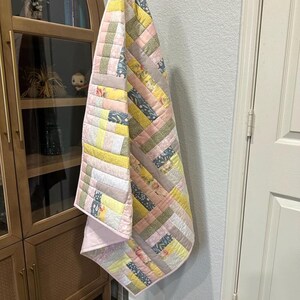 May include: A handmade quilt with a patchwork design. The quilt features a variety of fabrics in pastel colors, including pink, yellow, and gray. The quilt is hanging on a metal stand. The quilt's design is a mix of stripes and squares.