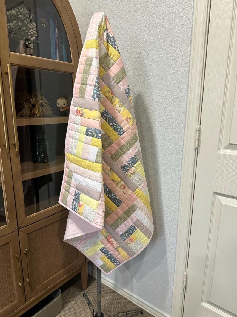 May include: A handmade quilt with a patchwork design. The quilt features a variety of fabrics in pastel colors, including pink, yellow, and gray. The quilt is hanging on a metal stand. The quilt's design is a mix of stripes and squares.