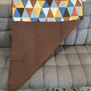 Puede incluir: Un edred&oacute;n marr&oacute;n y multicolor con un patr&oacute;n geom&eacute;trico de tri&aacute;ngulos. La secci&oacute;n superior presenta tri&aacute;ngulos en amarillo, azul, blanco y marr&oacute;n. La secci&oacute;n inferior es marr&oacute;n liso con un patr&oacute;n de puntos. El edred&oacute;n est&aacute; sobre un sof&aacute; gris.