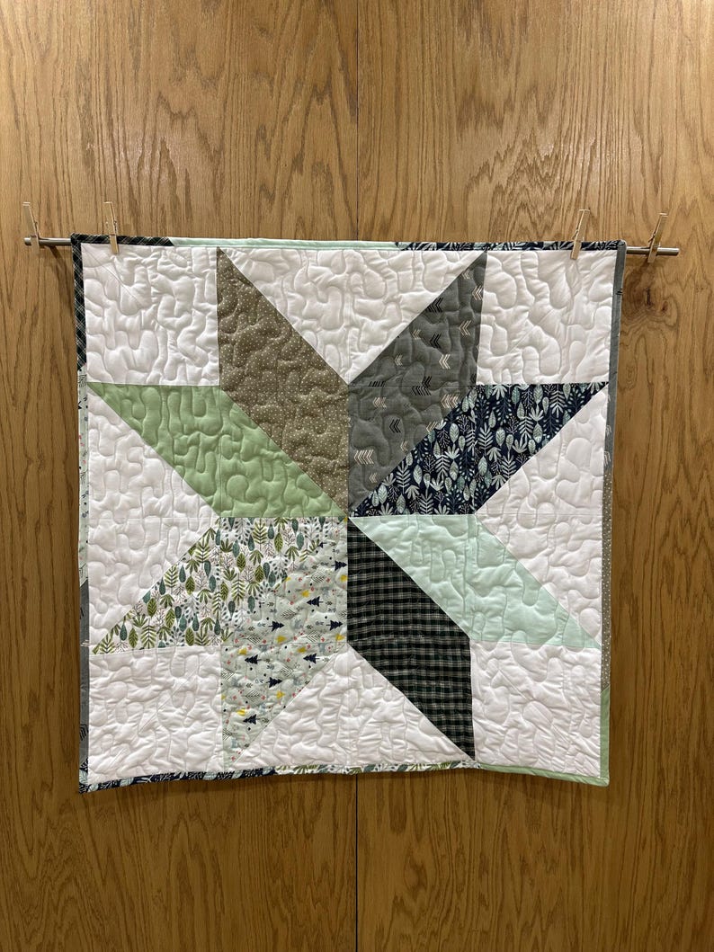 May include: A quilted star wall hanging with a variety of fabrics in shades of green, gray, and white. The star is centered on a white background with quilted texture. The quilt is hung on a wooden dowel with clothespins.