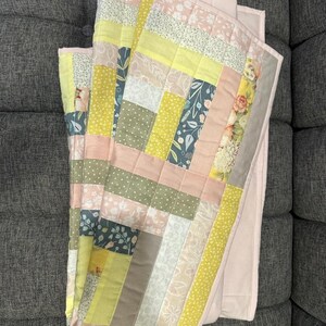 May include: A folded patchwork quilt with a variety of fabrics in pastel colors. The quilt features rectangular blocks of pink, yellow, green, and floral patterns. The quilt is folded on a gray surface.