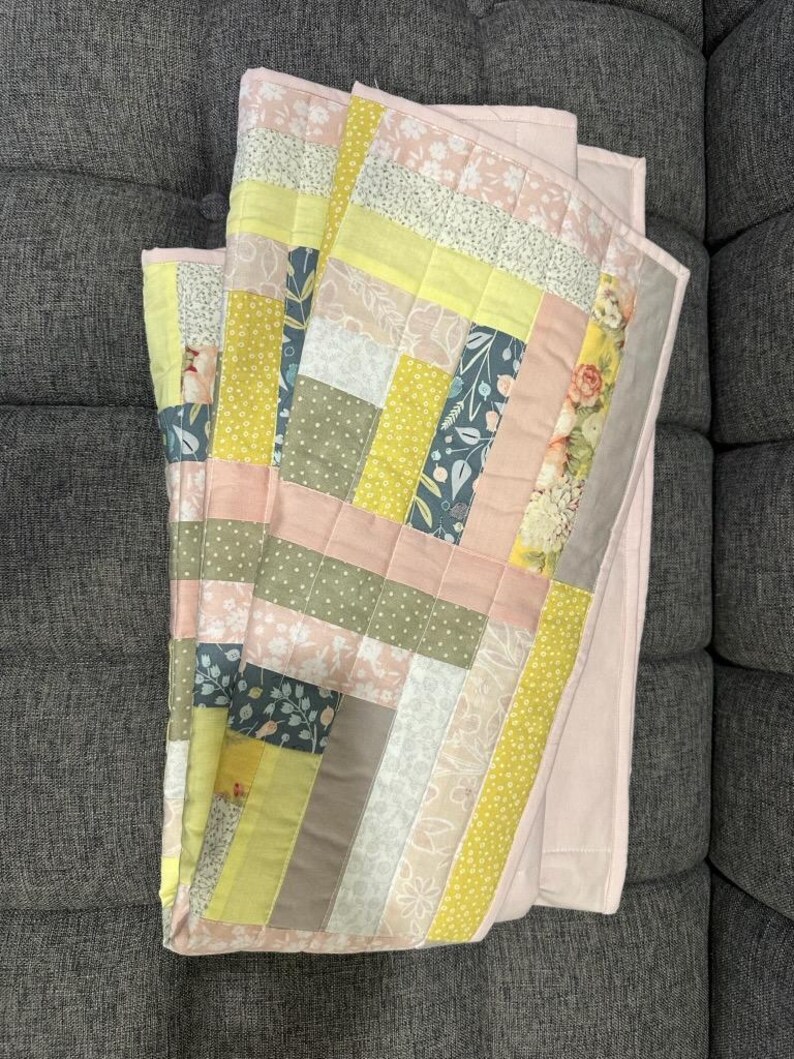 May include: A folded patchwork quilt with a variety of fabrics in pastel colors. The quilt features rectangular blocks of pink, yellow, green, and floral patterns. The quilt is folded on a gray surface.