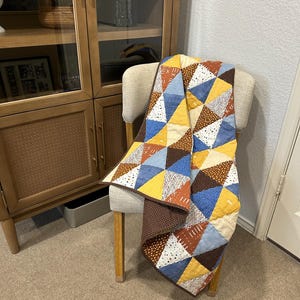 Puede incluir: Una colcha de retazos colorida sobre una silla. La colcha presenta un patr&oacute;n geom&eacute;trico de tri&aacute;ngulos en tonos de amarillo, azul, marr&oacute;n y blanco. La silla tiene un asiento beige claro y patas de madera. Un mueble de madera est&aacute; en el fondo.