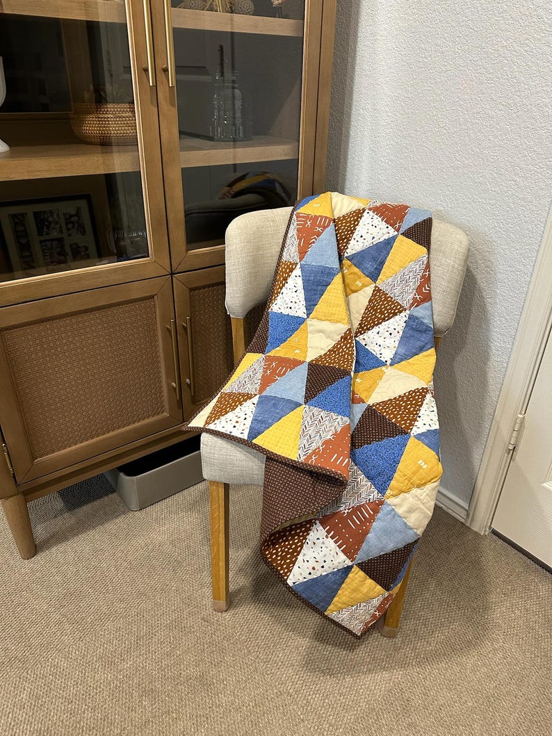 Puede incluir: Una colcha de retazos colorida sobre una silla. La colcha presenta un patr&oacute;n geom&eacute;trico de tri&aacute;ngulos en tonos de amarillo, azul, marr&oacute;n y blanco. La silla tiene un asiento beige claro y patas de madera. Un mueble de madera est&aacute; en el fondo.