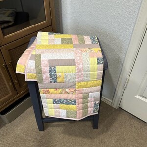 May include: A handmade patchwork quilt in pastel colors, including yellow, pink, and green. The quilt is draped over a dark blue side table. The quilt features a variety of floral and geometric patterns, creating a cozy and inviting aesthetic.