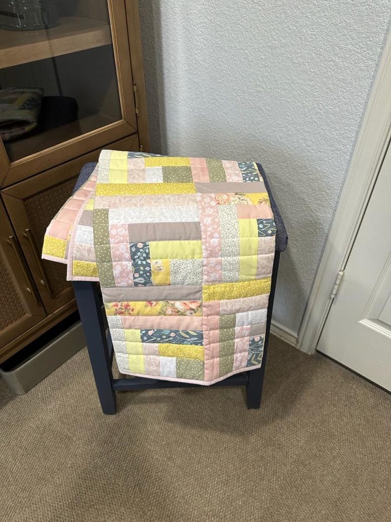 May include: A handmade patchwork quilt in pastel colors, including yellow, pink, and green. The quilt is draped over a dark blue side table. The quilt features a variety of floral and geometric patterns, creating a cozy and inviting aesthetic.