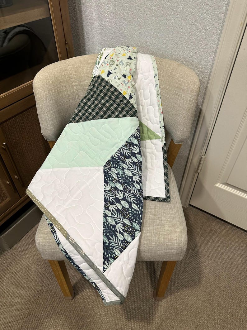 May include: A patchwork quilt draped over a beige chair. The quilt features a variety of patterns, including a black and green plaid, a mint green geometric design, and a navy blue leaf print. The quilt is trimmed with a gray border and white backing.