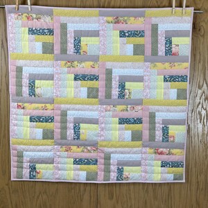 May include: A handmade quilt with a log cabin design. The quilt features squares of fabric in various colors, including pink, yellow, gray, and floral patterns. It is hanging on a clothesline with wooden clothespins.