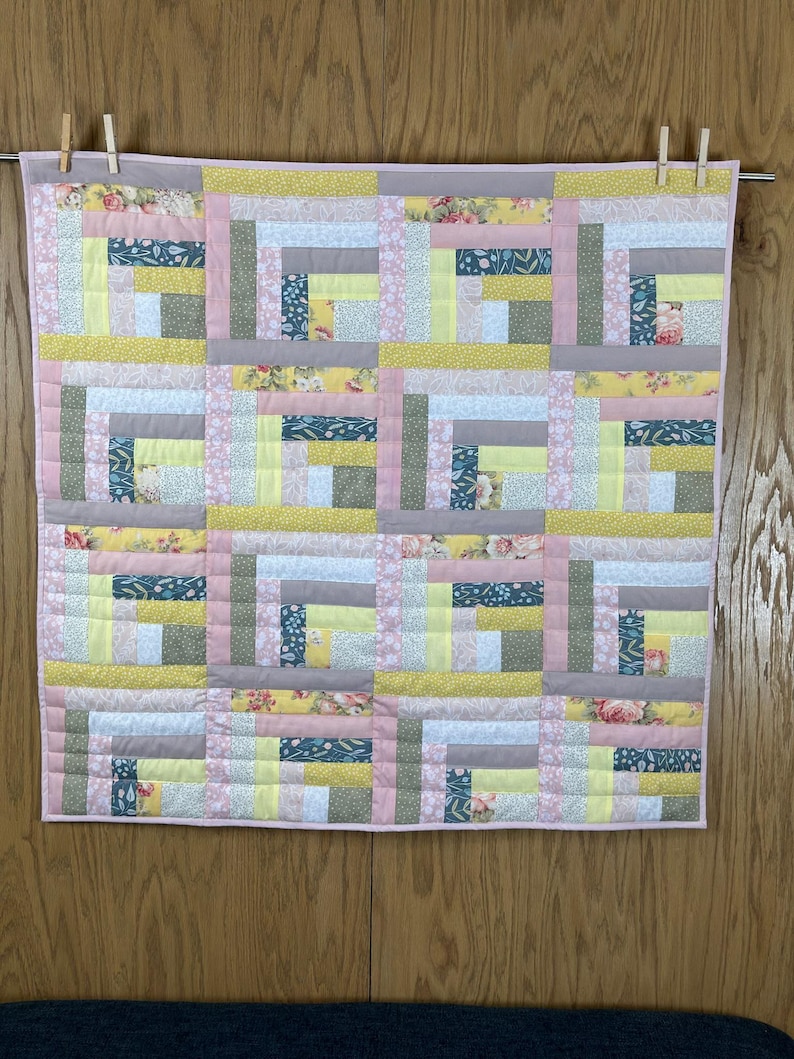 May include: A handmade quilt with a log cabin design. The quilt features squares of fabric in various colors, including pink, yellow, gray, and floral patterns. It is hanging on a clothesline with wooden clothespins.