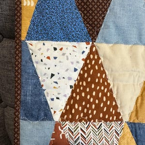 Puede incluir: Primer plano de un edred&oacute;n hecho a mano con un dise&ntilde;o de patchwork. El edred&oacute;n presenta varias formas geom&eacute;tricas en tonos de azul, marr&oacute;n, amarillo y blanco. Las telas incluyen textiles florales, punteados y estampados, cosidos con un borde decorativo.