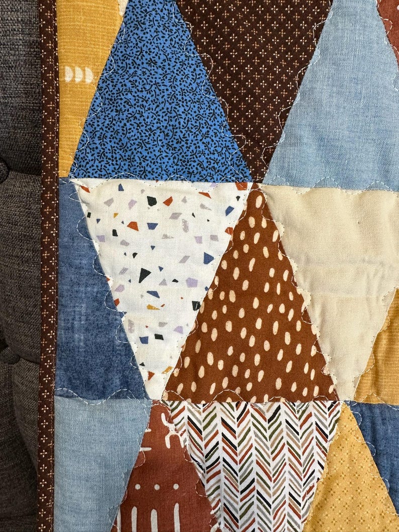 Puede incluir: Primer plano de un edred&oacute;n hecho a mano con un dise&ntilde;o de patchwork. El edred&oacute;n presenta varias formas geom&eacute;tricas en tonos de azul, marr&oacute;n, amarillo y blanco. Las telas incluyen textiles florales, punteados y estampados, cosidos con un borde decorativo.