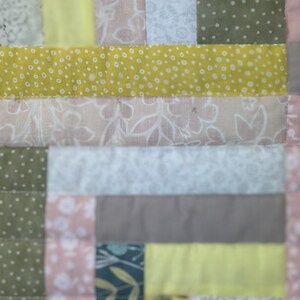 May include: Close-up of a patchwork quilt with a variety of fabrics. The quilt features squares and rectangles in shades of yellow, pink, green, and gray. Some fabrics have floral or dotted patterns, while others are solid colors. The quilt is stitched together with a visible seam.