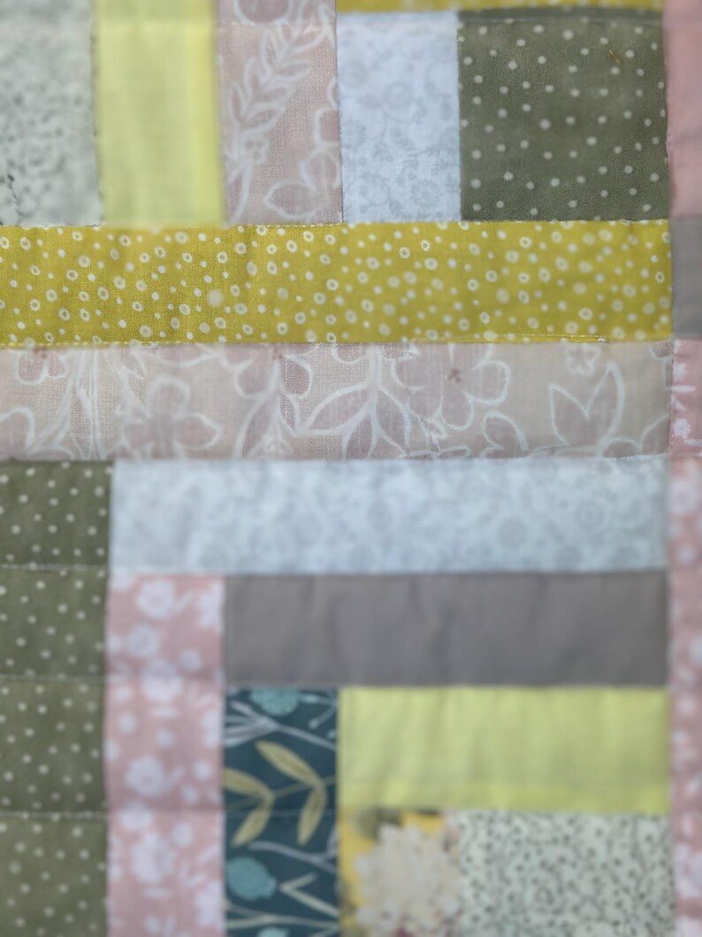 May include: Close-up of a patchwork quilt with a variety of fabrics. The quilt features squares and rectangles in shades of yellow, pink, green, and gray. Some fabrics have floral or dotted patterns, while others are solid colors. The quilt is stitched together with a visible seam.