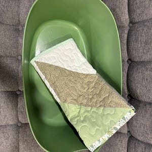 May include: A green plastic laundry basket holds a small, quilted baby blanket. The blanket features a white section, a section with small brown polka dots, and a light green section. The blanket has a floral patterned border.