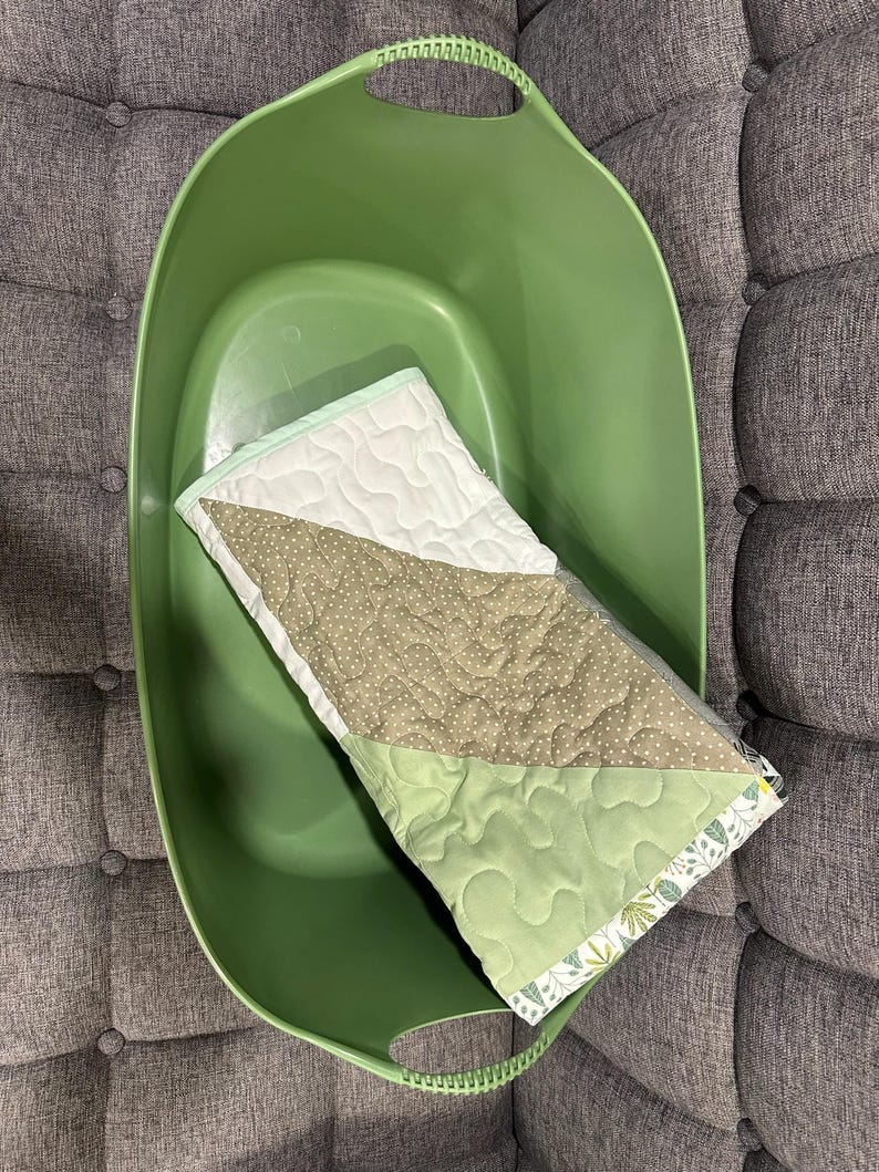 May include: A green plastic laundry basket holds a small, quilted baby blanket. The blanket features a white section, a section with small brown polka dots, and a light green section. The blanket has a floral patterned border.