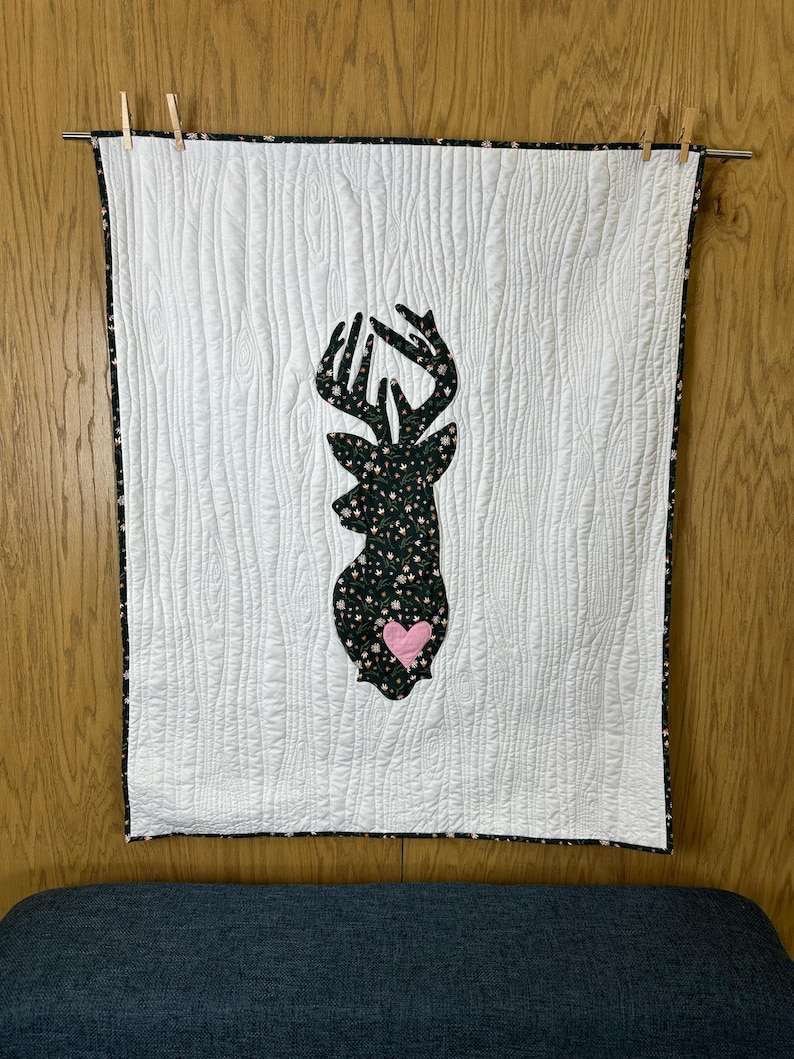 May include: White quilted wall hanging with a deer silhouette. The deer is filled with a floral pattern and a pink heart. The edges are trimmed with a floral pattern. The wall hanging is hung on a metal rod with wooden clothespins.