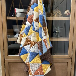Puede incluir: Un edred&oacute;n hecho a mano con un dise&ntilde;o de retazos. El edred&oacute;n presenta tri&aacute;ngulos en tonos de azul, amarillo, marr&oacute;n y blanco. El edred&oacute;n est&aacute; colgado en un armario de madera. Sus dimensiones son de aproximadamente 152 cm por 152 cm.