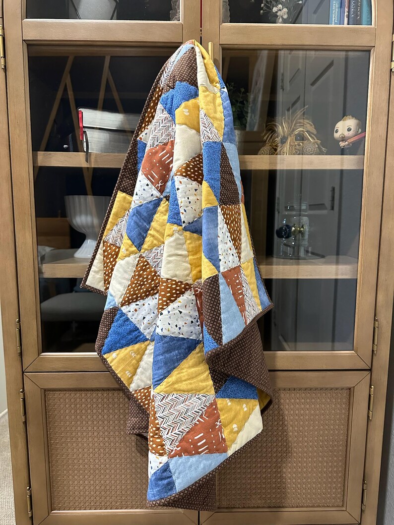 Puede incluir: Un edred&oacute;n hecho a mano con un dise&ntilde;o de retazos. El edred&oacute;n presenta tri&aacute;ngulos en tonos de azul, amarillo, marr&oacute;n y blanco. El edred&oacute;n est&aacute; colgado en un armario de madera. Sus dimensiones son de aproximadamente 152 cm por 152 cm.