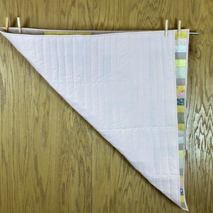 May include: A light pink and white quilt, partially displayed, hanging from a metal rod with wooden clothespins. The quilt has vertical stitching and a colorful patterned border with shades of green, yellow, and blue.