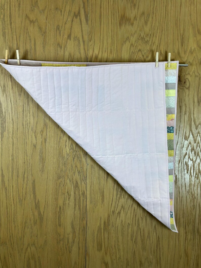 May include: A light pink and white quilt, partially displayed, hanging from a metal rod with wooden clothespins. The quilt has vertical stitching and a colorful patterned border with shades of green, yellow, and blue.