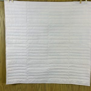 May include: A light pink, quilted blanket hanging from a metal rod with two wooden clothespins. The blanket has horizontal stitching creating a ribbed texture. The background is a wooden wall.