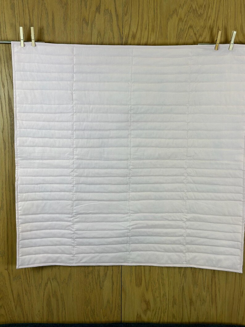 May include: A light pink, quilted blanket hanging from a metal rod with two wooden clothespins. The blanket has horizontal stitching creating a ribbed texture. The background is a wooden wall.