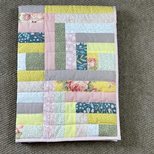 May include: A patchwork quilt with a variety of floral and geometric patterns in shades of pink, yellow, gray, and blue. The quilt has a pink border and is likely handmade. The quilt is rectangular and appears to be a baby quilt or lap blanket.