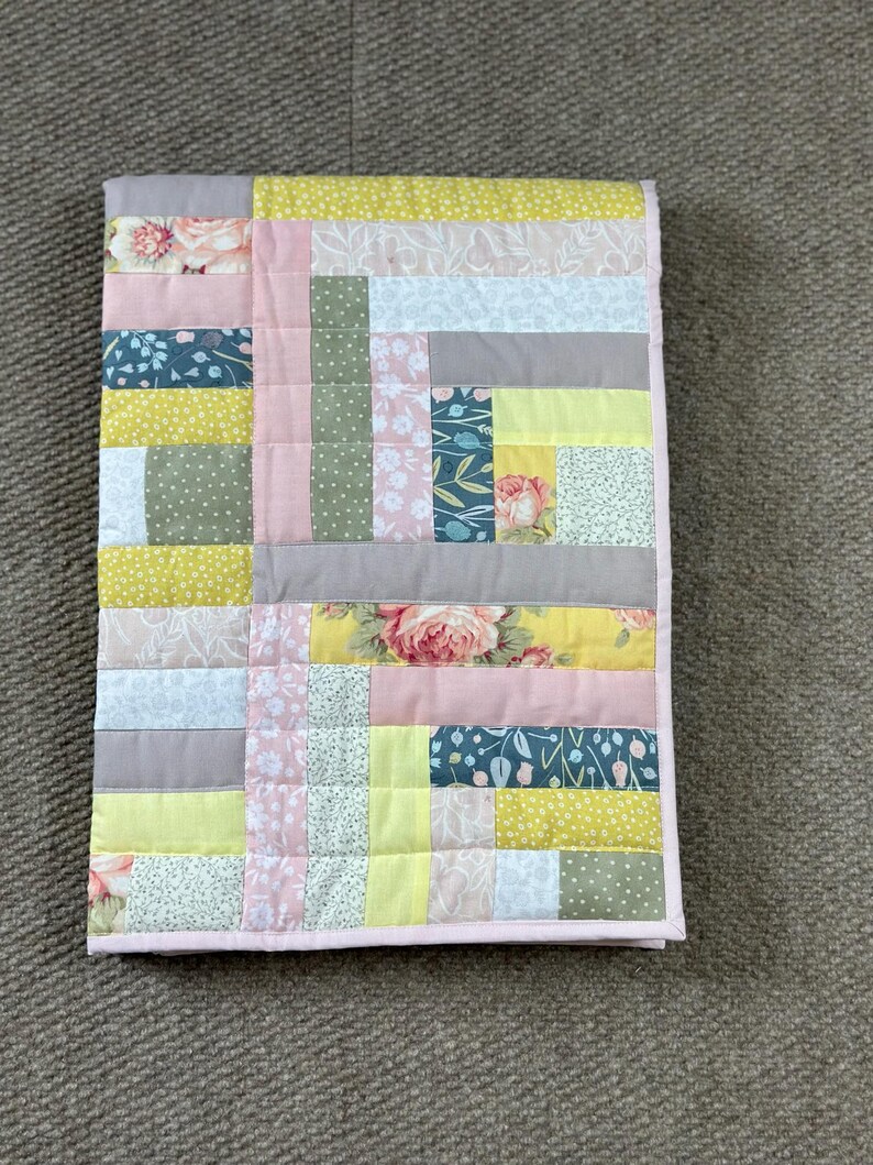 May include: A patchwork quilt with a variety of floral and geometric patterns in shades of pink, yellow, gray, and blue. The quilt has a pink border and is likely handmade. The quilt is rectangular and appears to be a baby quilt or lap blanket.