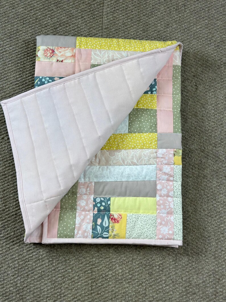 May include: A patchwork quilt with a light pink backing and a variety of patterned squares in pink, green, yellow, and gray. The quilt is folded over, revealing the quilted backing.