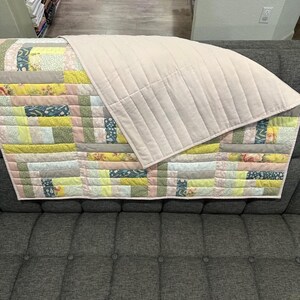 May include: A handmade quilt with a patchwork design in pastel colors, including pink, yellow, and green. The quilt is draped over a gray sofa. The top layer is a solid, light pink fabric with vertical stitching.