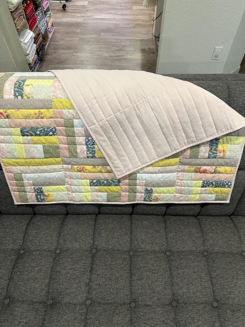 May include: A handmade quilt with a patchwork design in pastel colors, including pink, yellow, and green. The quilt is draped over a gray sofa. The top layer is a solid, light pink fabric with vertical stitching.
