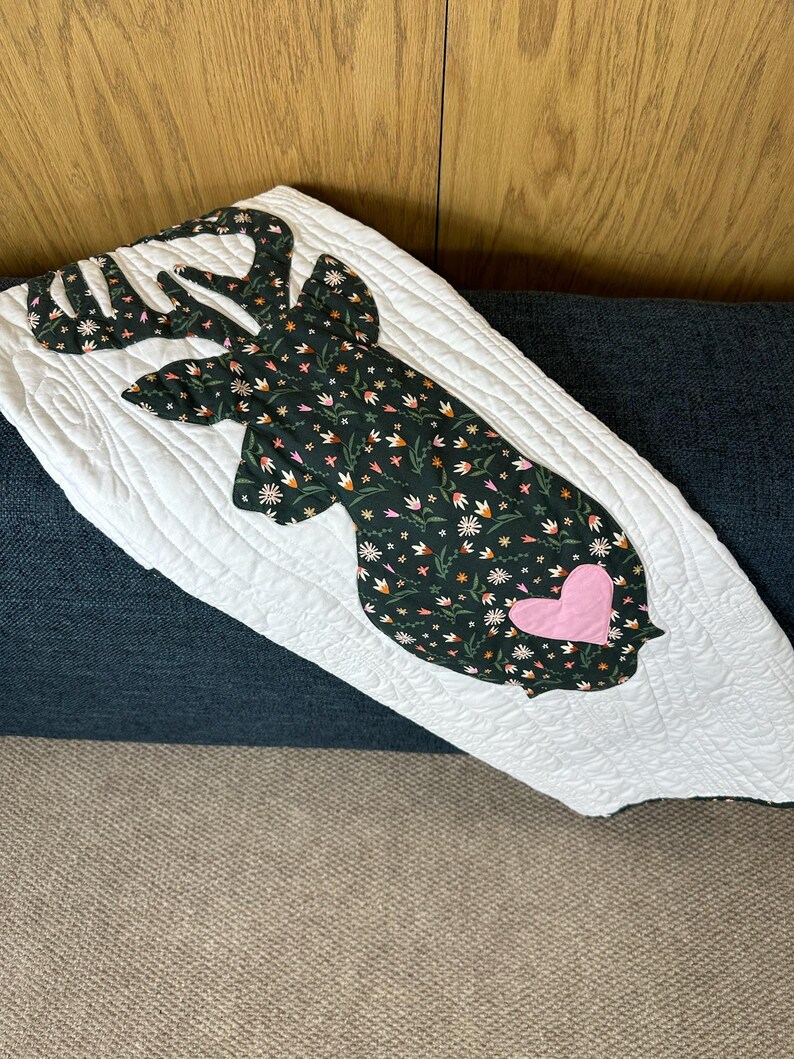 May include: White quilted throw blanket featuring a deer head silhouette. The deer head is made of dark green fabric with a floral pattern and a pink heart. The blanket is on a blue sofa.