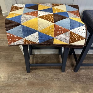 Puede incluir: Una funda de taburete de patchwork hecha a mano con un dise&ntilde;o geom&eacute;trico. La funda est&aacute; hecha de varias telas en tonos de azul, amarillo, marr&oacute;n y blanco. La funda est&aacute; colocada sobre un taburete gris oscuro.
