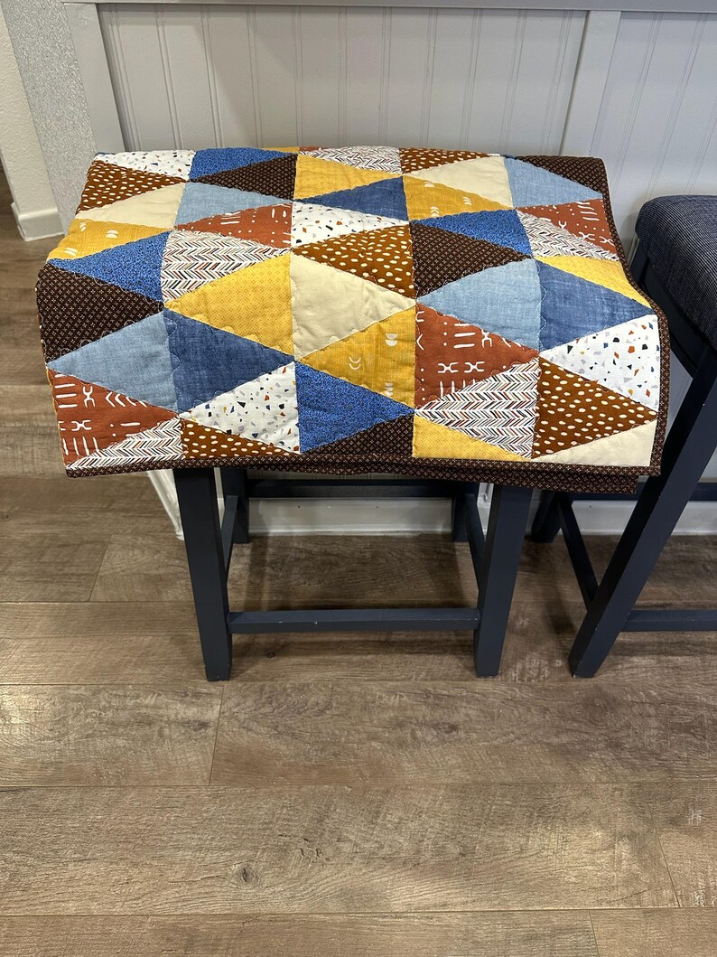 Puede incluir: Una funda de taburete de patchwork hecha a mano con un dise&ntilde;o geom&eacute;trico. La funda est&aacute; hecha de varias telas en tonos de azul, amarillo, marr&oacute;n y blanco. La funda est&aacute; colocada sobre un taburete gris oscuro.