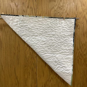 May include: A white quilted blanket with a wavy pattern is displayed, hanging from a wooden rod with clothespins. The blanket has a patterned border with shades of blue, green, and white. The background is a wooden surface.