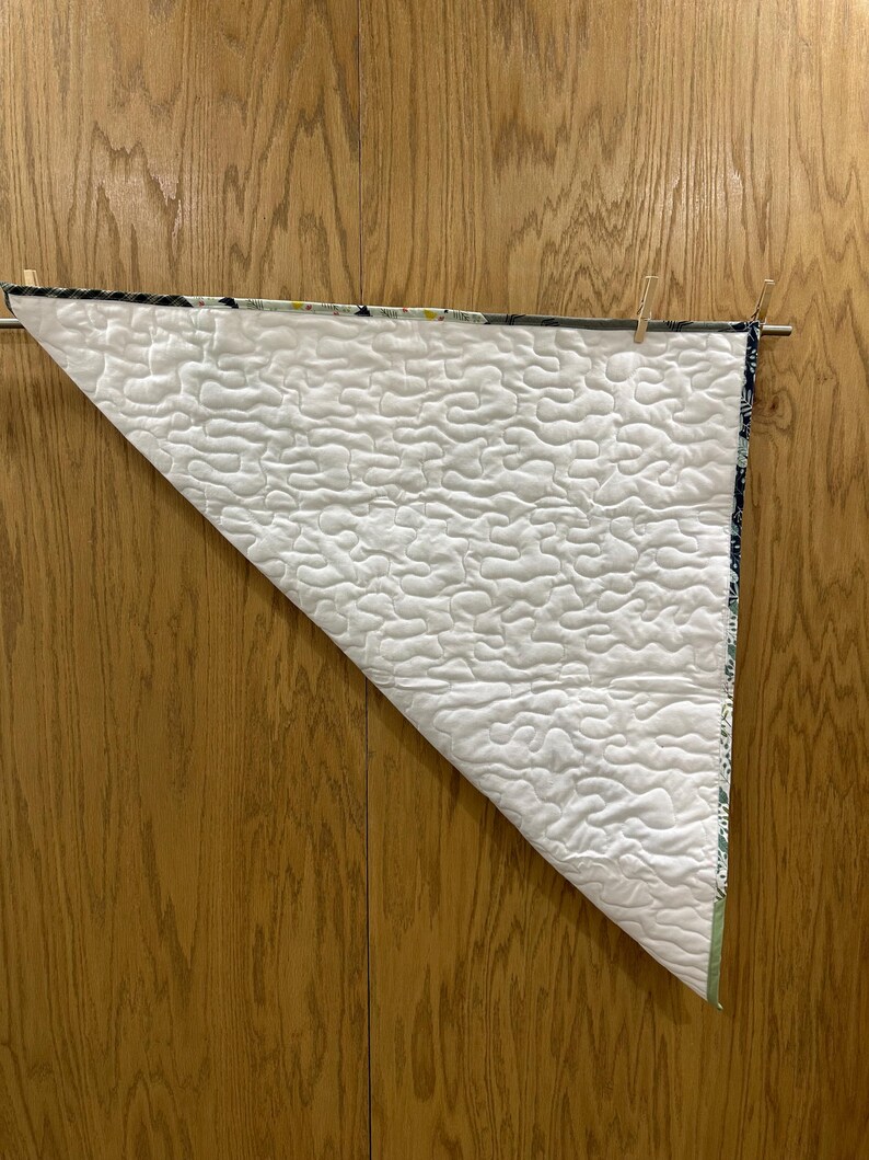 May include: A white quilted blanket with a wavy pattern is displayed, hanging from a wooden rod with clothespins. The blanket has a patterned border with shades of blue, green, and white. The background is a wooden surface.