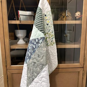 May include: A patchwork quilt with a variety of patterned fabrics, including green plaid, floral, and geometric designs. The quilt features a white quilted border and a light blue binding. The quilt is hanging on a wooden cabinet.