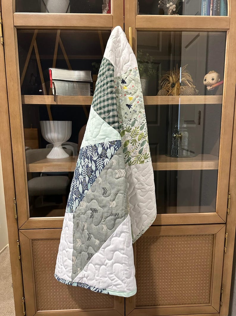 May include: A patchwork quilt with a variety of patterned fabrics, including green plaid, floral, and geometric designs. The quilt features a white quilted border and a light blue binding. The quilt is hanging on a wooden cabinet.